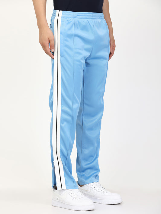 A person standing wearing blue side tape straight fit track pants with white stripes on the side and a pair of white sneakers.