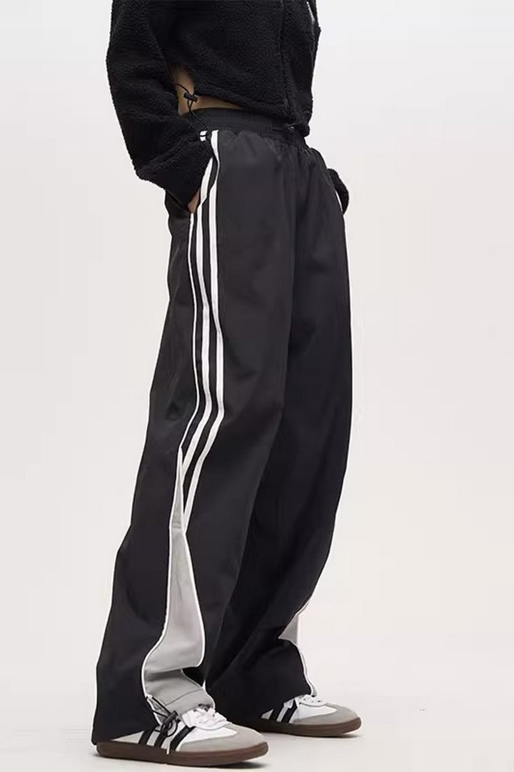 A person standing sideways, wearing black striped parachute pants with white contrast piping, an elasticated waistband with adjustable drawstrings, and white sneakers.
