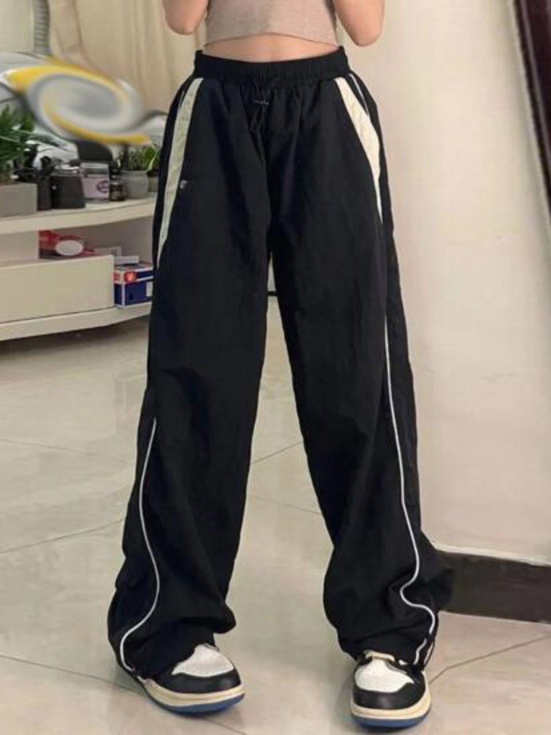 A pair of black track pants with contrasting white piping on the sides, worn by a person standing against a white background.