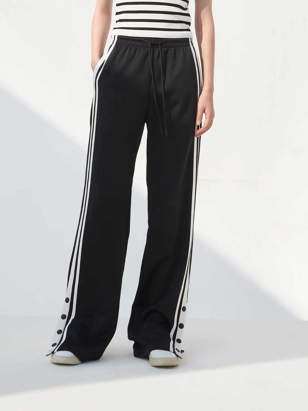 A pair of black slit snap button tracks pants with an elastic drawstring waist, featuring white stripes on the legs and a side slit on one leg. The model is wearing a white top and beige shoes.