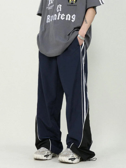 A person standing side-on, wearing striped parachute pants with contrast piping and an elasticated waistband with adjustable drawstrings.