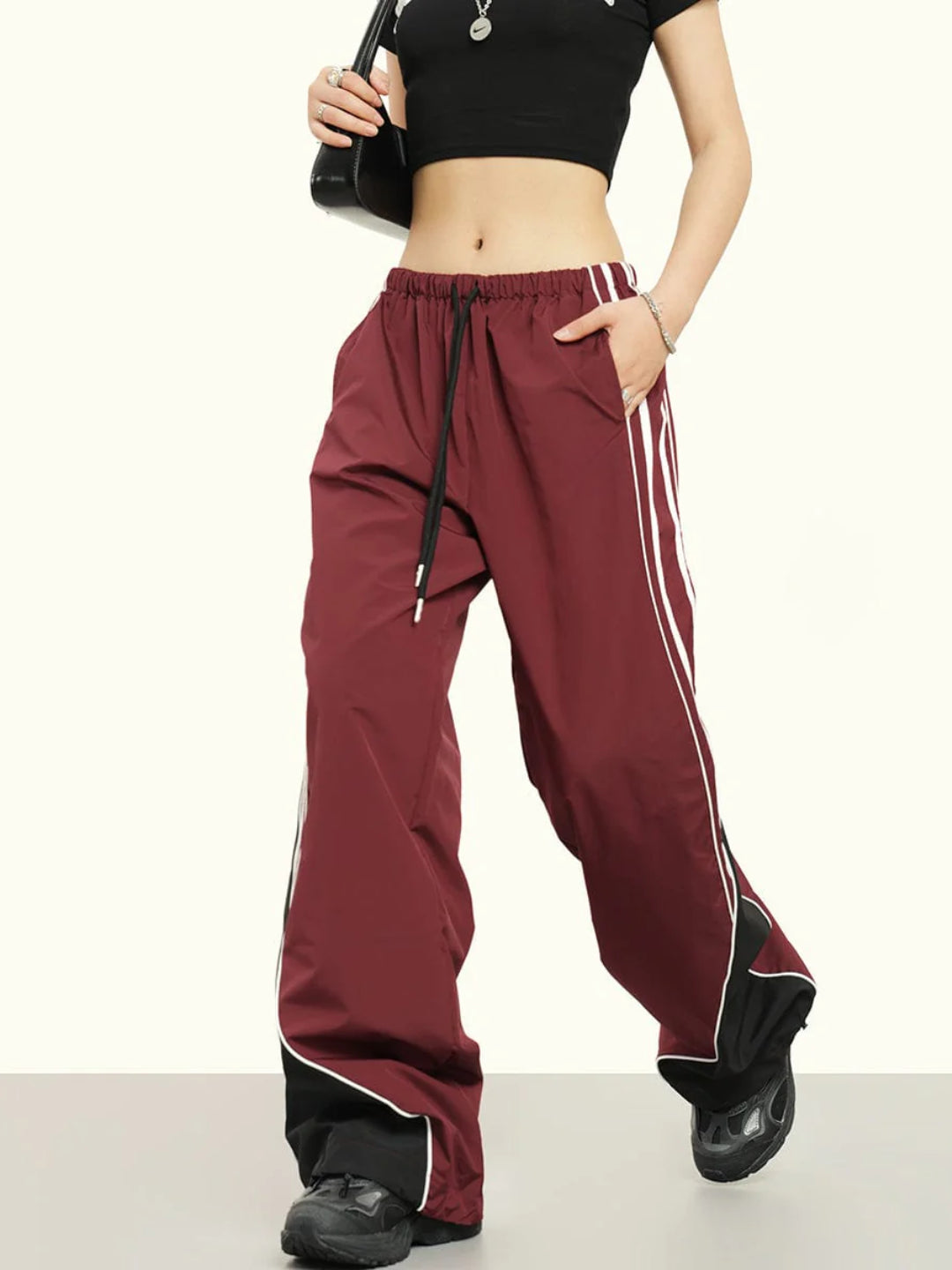 A person wearing striped parachute pants with contrast piping, an elasticated waistband with drawstrings, in a maroon color.