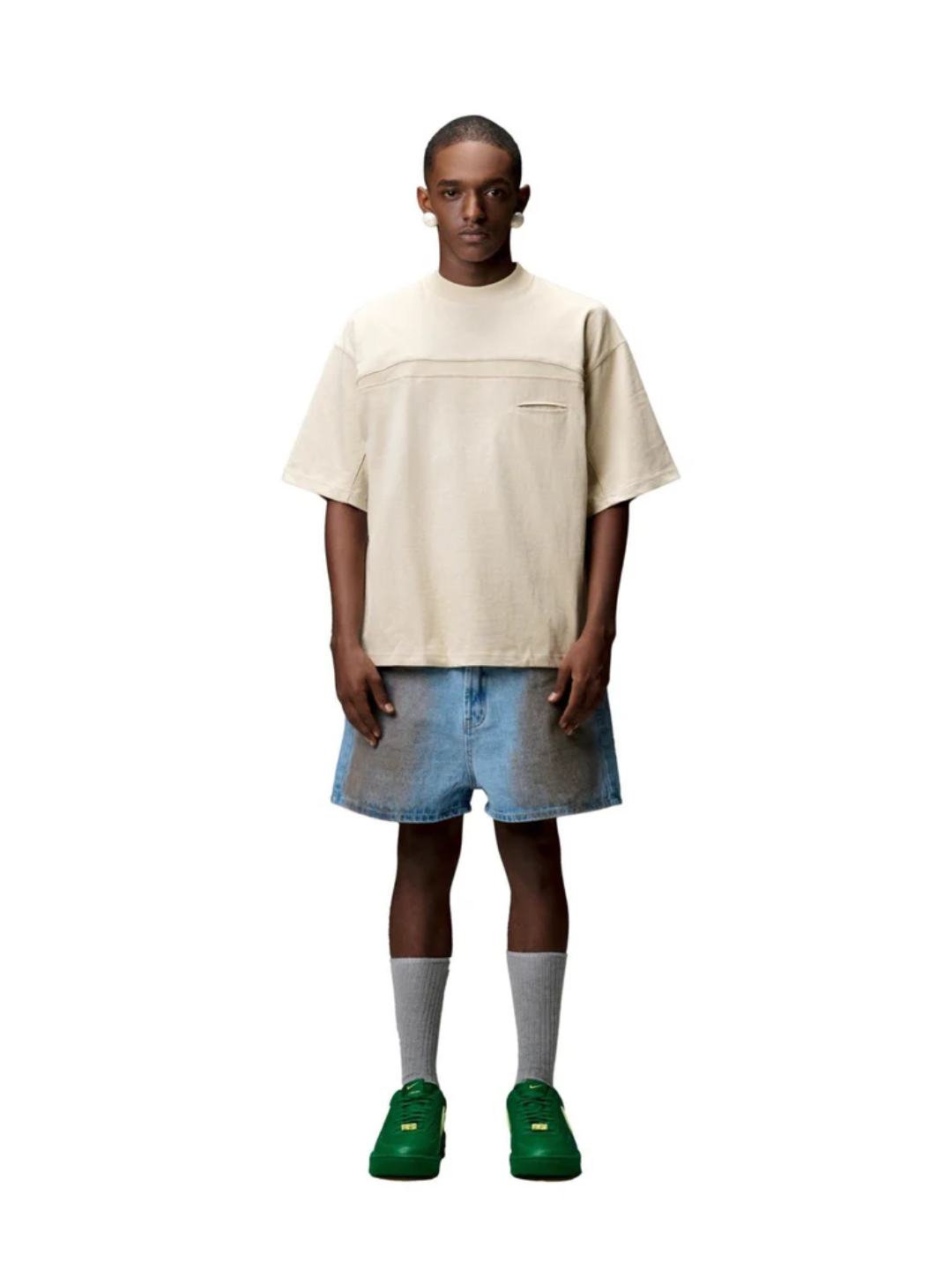 A person wearing a beige Boxy Fit tee, blue denim shorts, green sneakers, and white socks standing against a gray background.