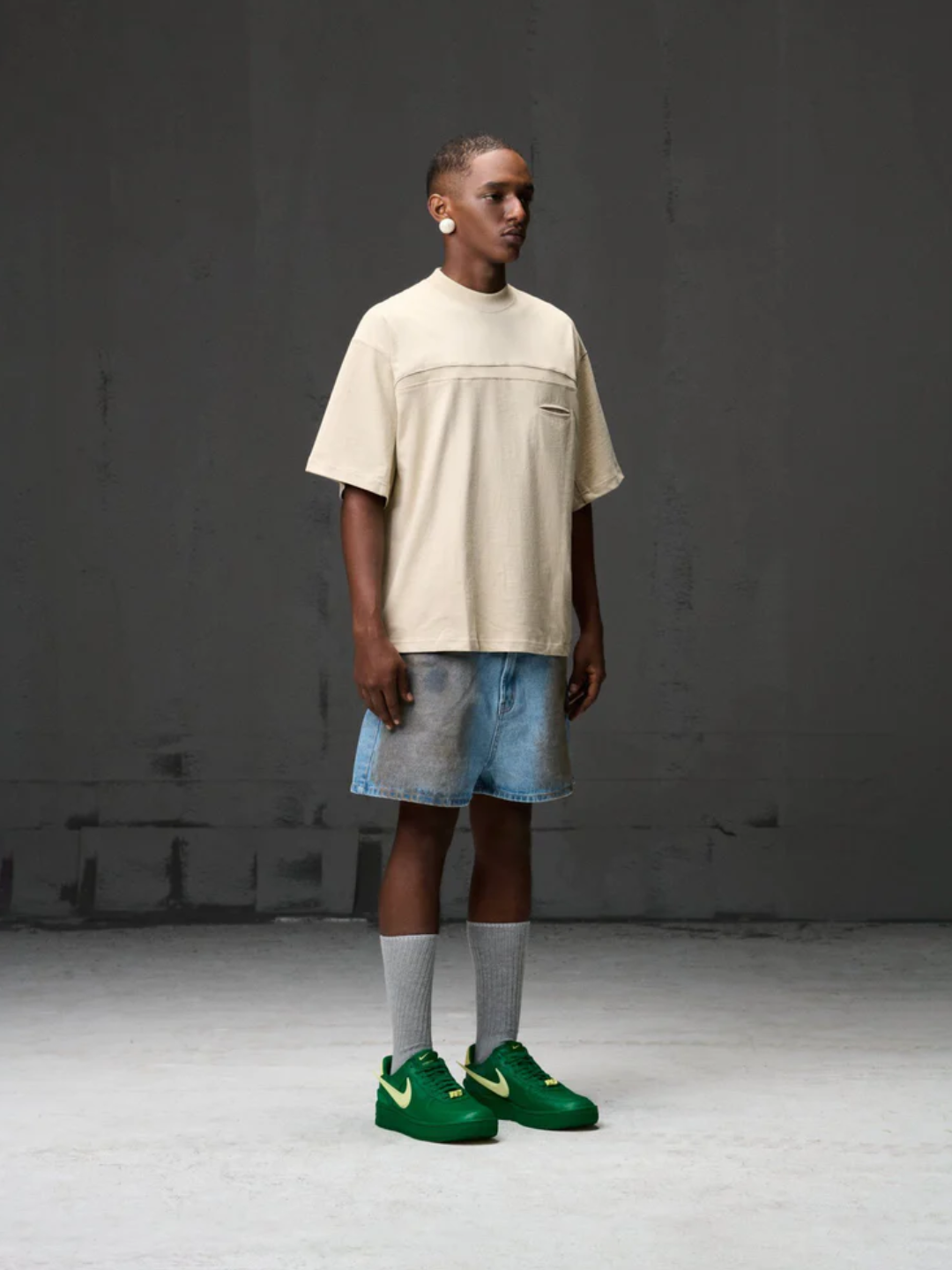 A male model wearing a beige boxy fit tee with a collar, short sleeves, and a front pocket, paired with grey shorts and green sneakers.