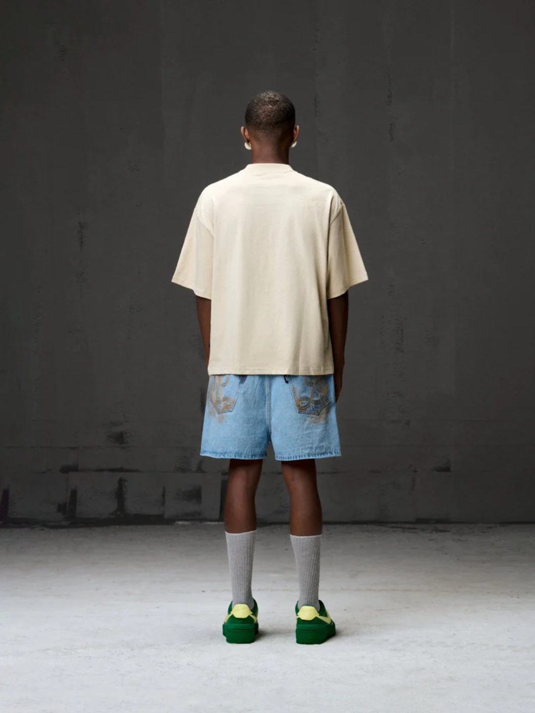 A person wearing a beige Boxy Fit tee, blue denim shorts, green sneakers, and white socks standing against a gray background.