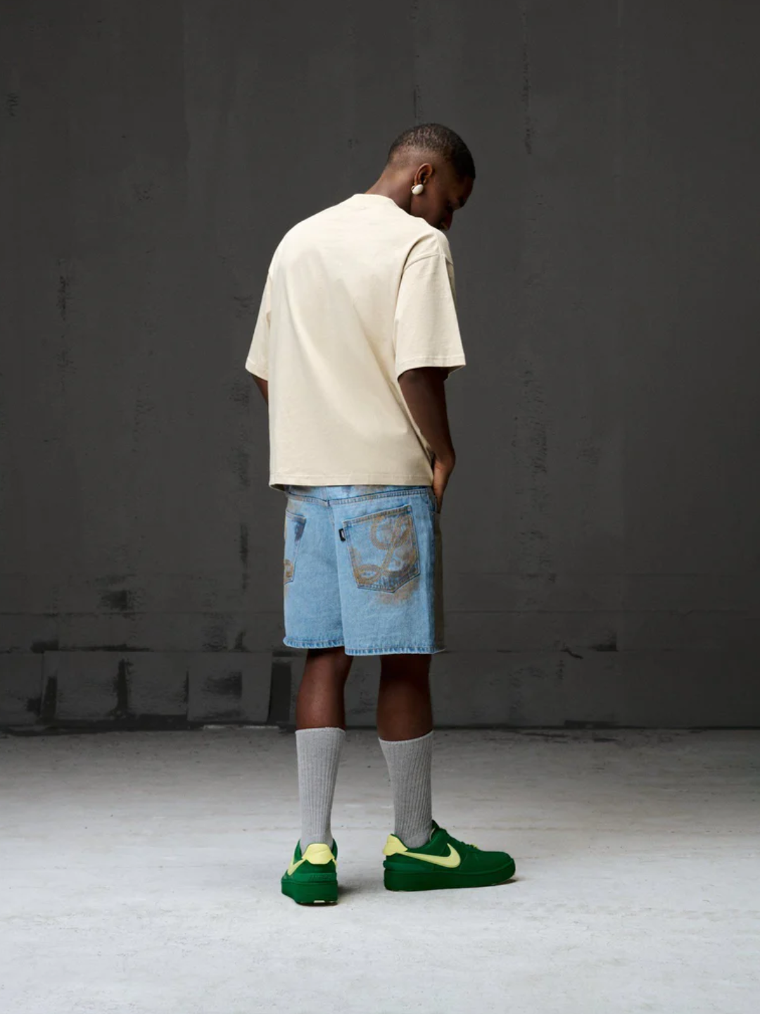 A person wearing a beige Boxy Fit tee, blue denim shorts, green sneakers, and white socks standing against a gray background.