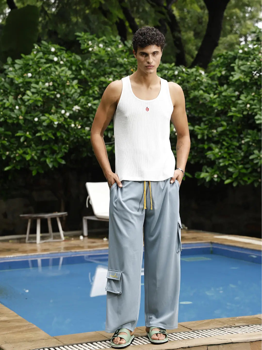 Man standing by a pool wearing a white tank top and light gray pants with cargo pockets.