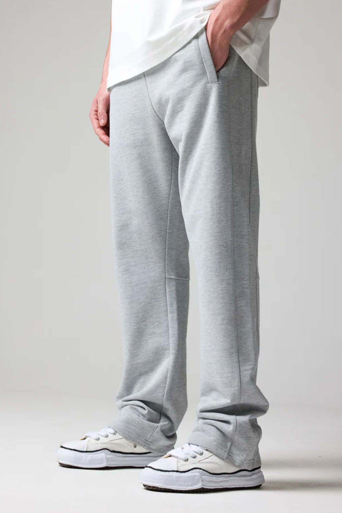 A person standing sideways, wearing grey open hem joggers with an elastic waistband and drawstring, along with a white t-shirt and white sneakers.