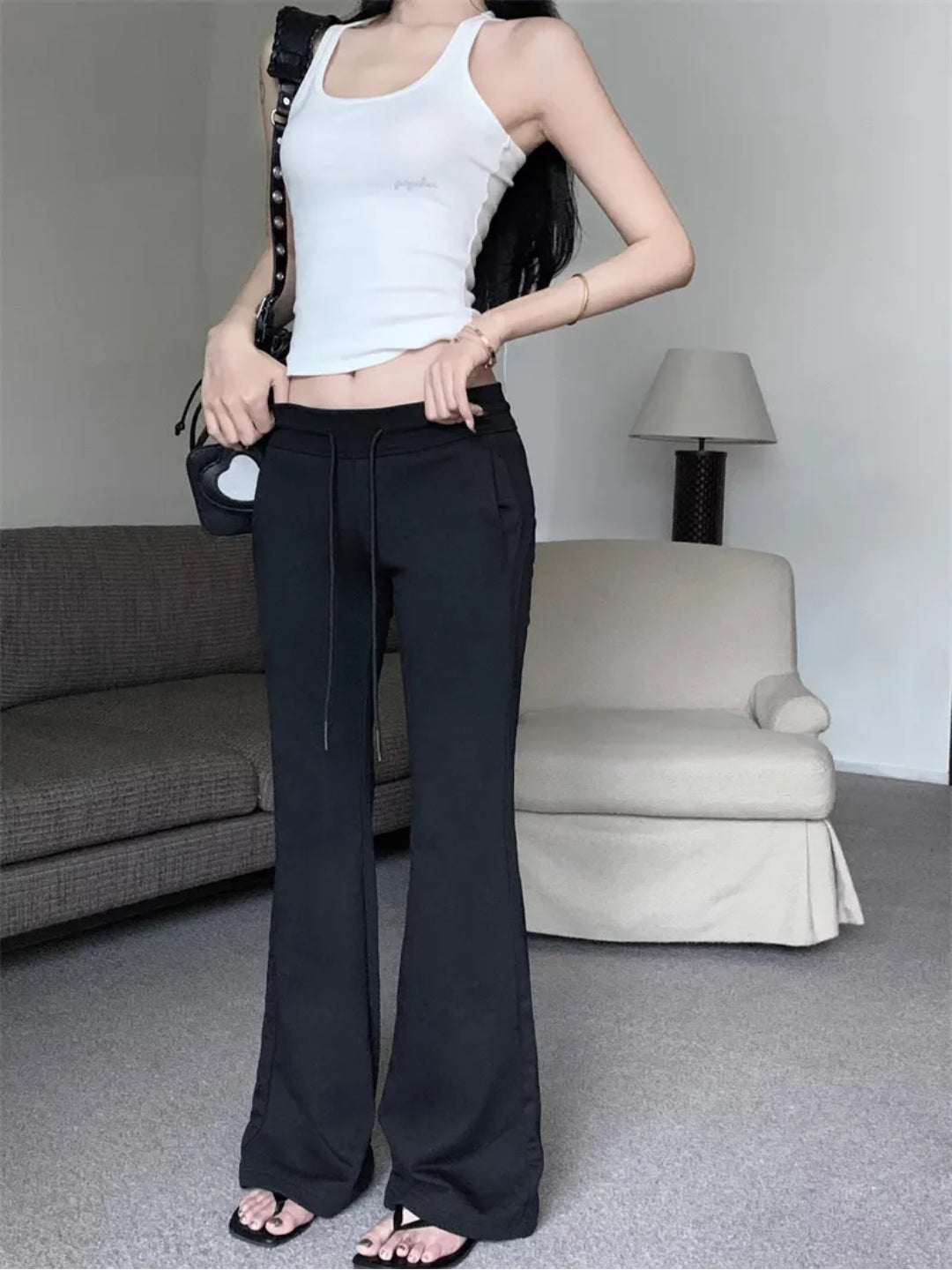 A person standing in a room with furniture, wearing black low rise flared sweatpants with a white tank top.