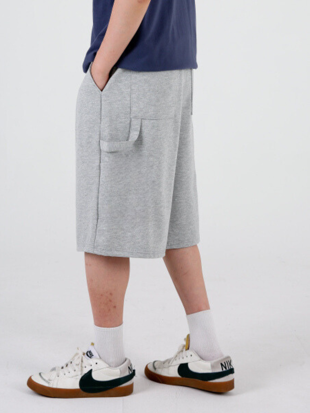 A person standing sideways, wearing gray carpenter shorts with an elastic waistband and drawstring, paired with a blue top and white sneakers.