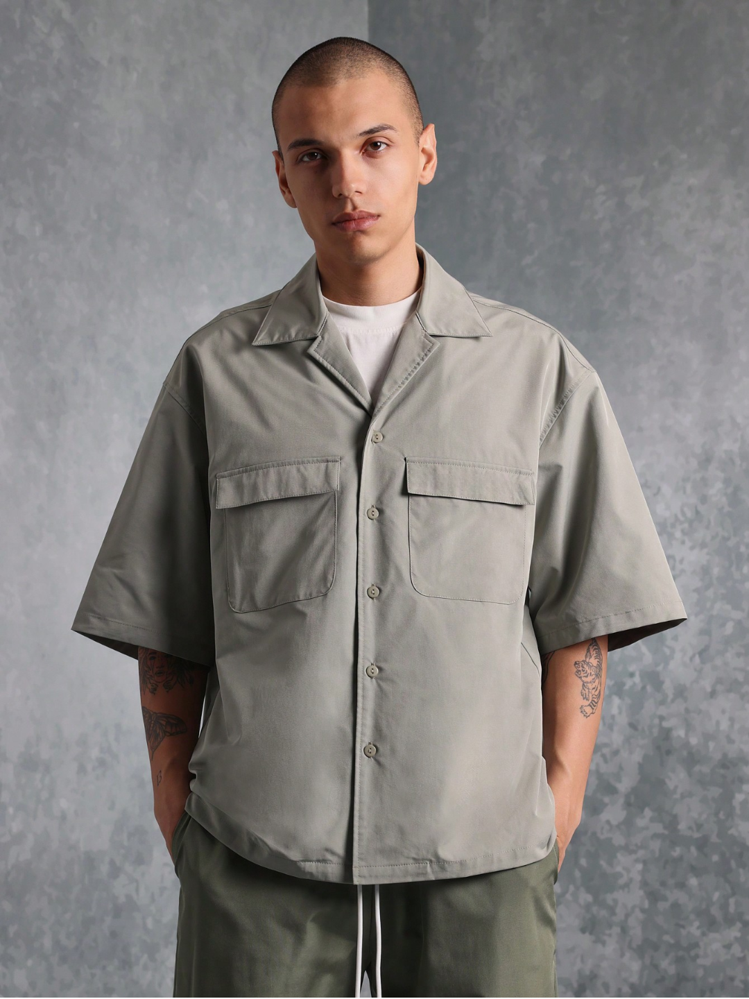 This image features the Chateau Boxy Fit Shirt and Pant set, presenting a stylish and relaxed outfit. The shirt is short-sleeved with a boxy fit and features two chest pockets, in a light grey color that complements the darker green pants.