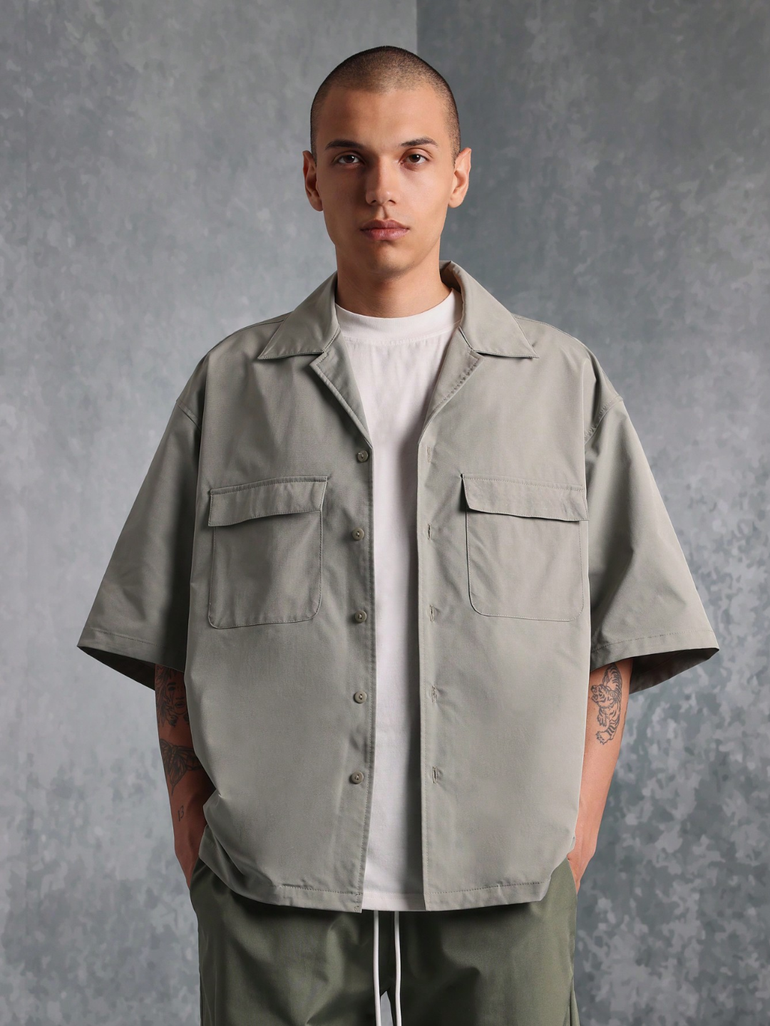 This image features the Chateau Boxy Fit Shirt and Pant set, presenting a stylish and relaxed outfit. The shirt is short-sleeved with a boxy fit and features two chest pockets, in a light grey color that complements the darker green pants.