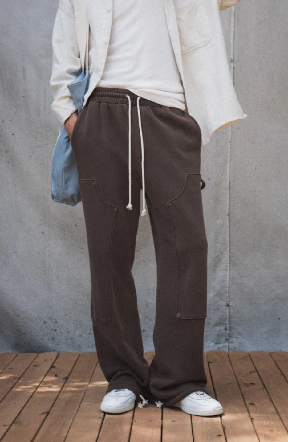 A person standing on a wooden surface wearing brown straight-leg carpenter pants with an elastic waistband and drawcord, paired with a white jacket and sneakers.