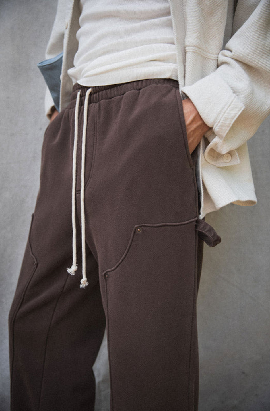 A person standing on a wooden surface wearing brown straight-leg carpenter pants with an elastic waistband and drawcord, paired with a white jacket and sneakers.