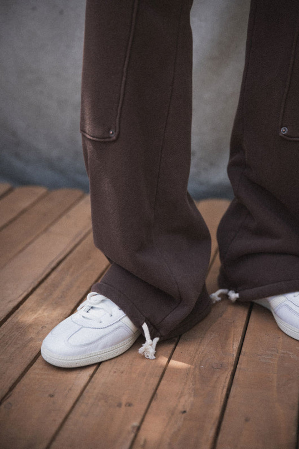 A person standing on a wooden surface wearing brown straight-leg carpenter pants with an elastic waistband and drawcord, paired with a white jacket and sneakers.