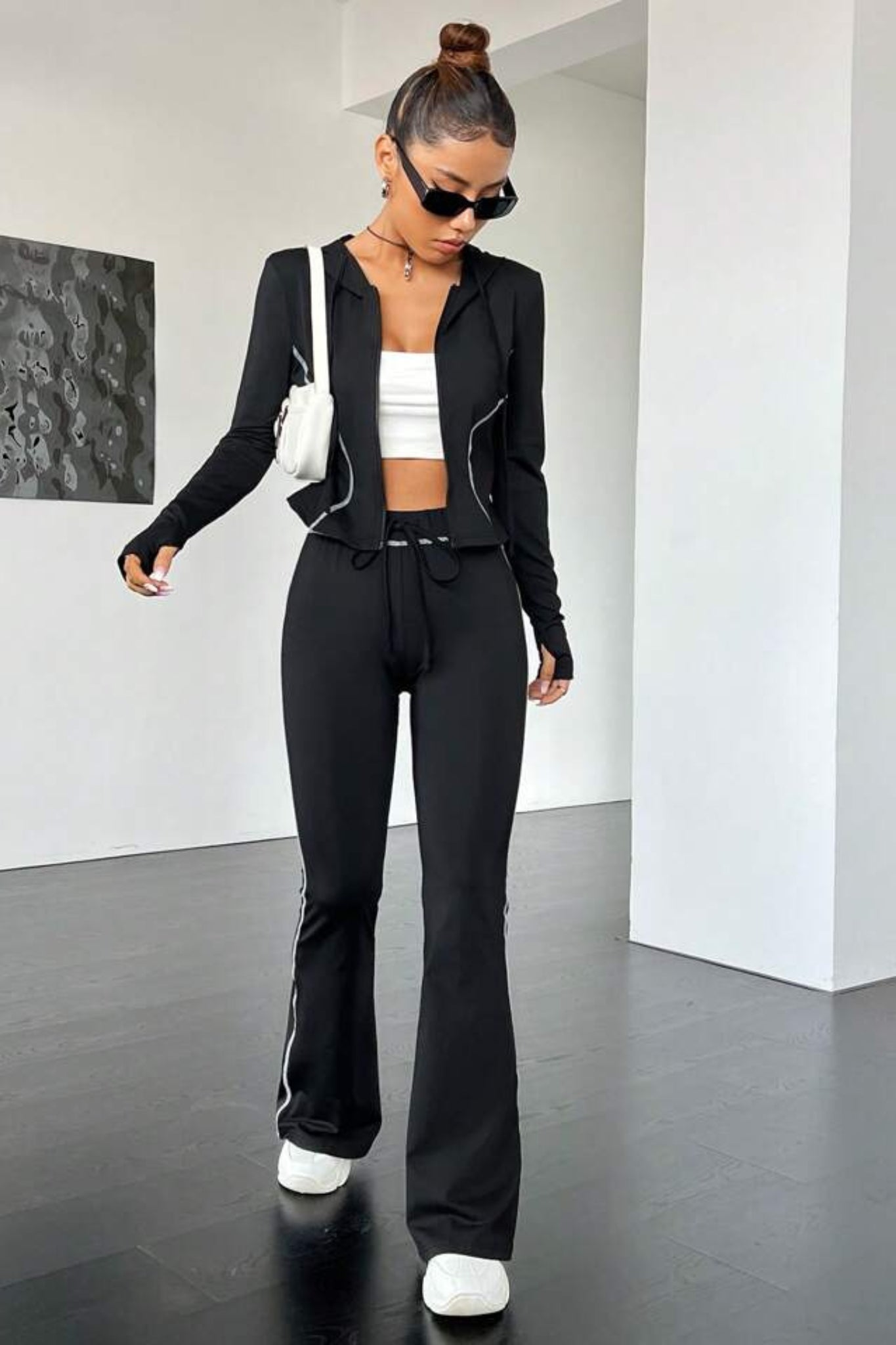 A woman wearing a black contrast stitch tracksuit with a zip-up jacket and matching leggings, accessorized with a white crop top and white sneakers.