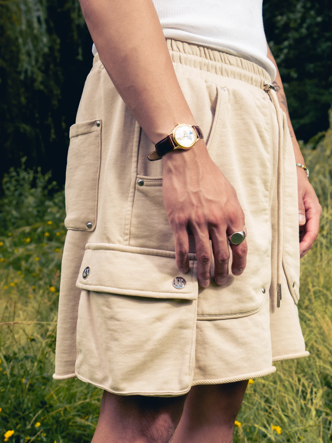 A pair of beige cargo sweat shorts with multiple pockets and a drawstring waistband, shown from the waist to the mid-thigh on a person standing in a field.