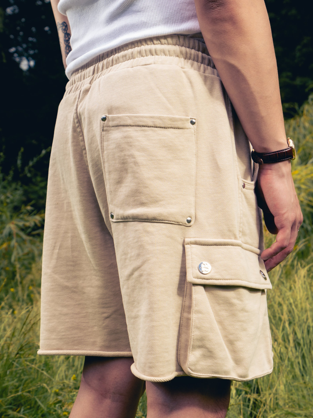 A pair of beige cargo sweat shorts with multiple pockets and a drawstring waistband, shown from the waist to the mid-thigh on a person standing in a field.
