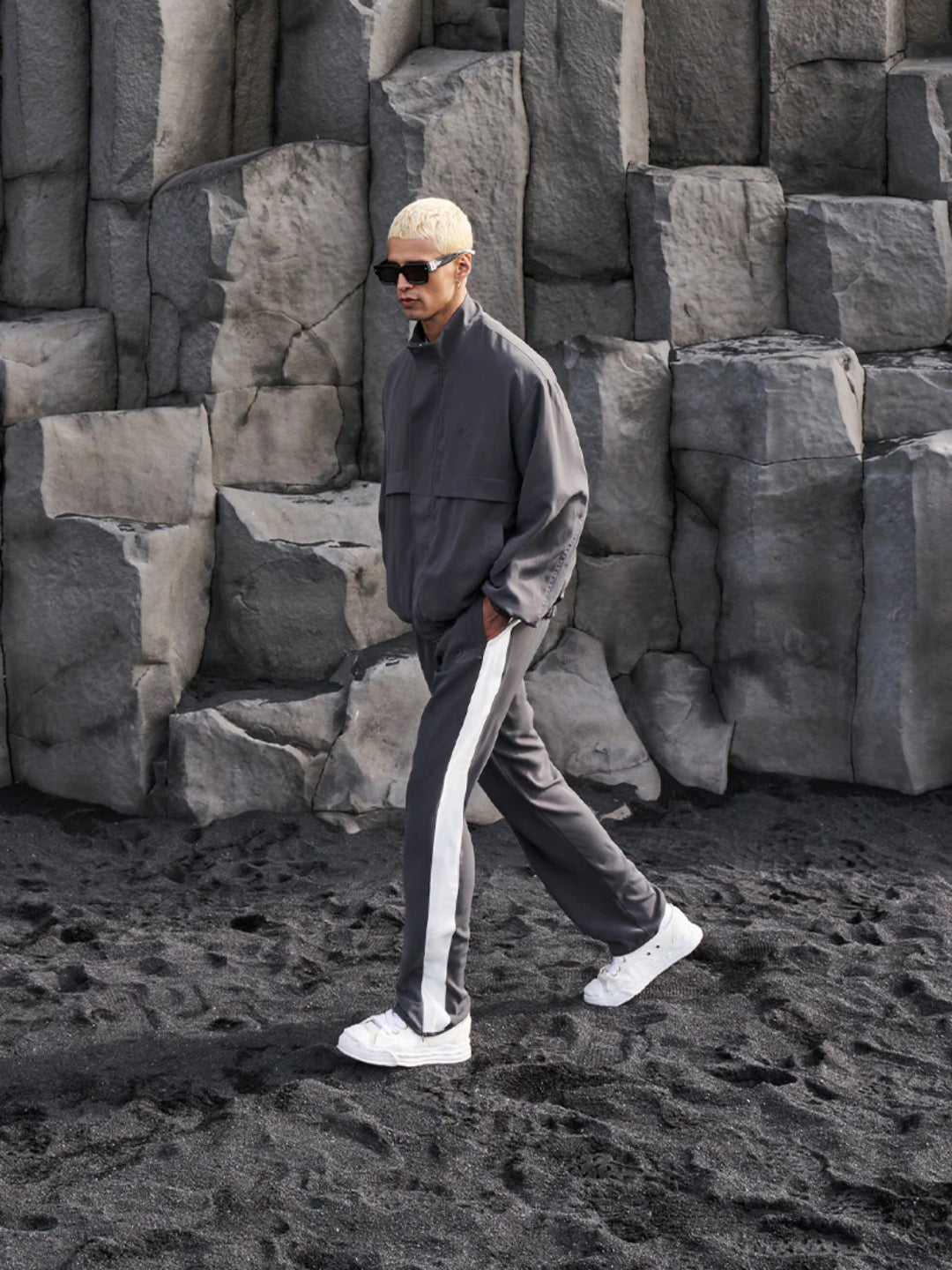 A man standing wearing a dark gray tracksuit, consisting of a jacket with a funnel neck and front pockets, and matching pants with an elasticated waistband.