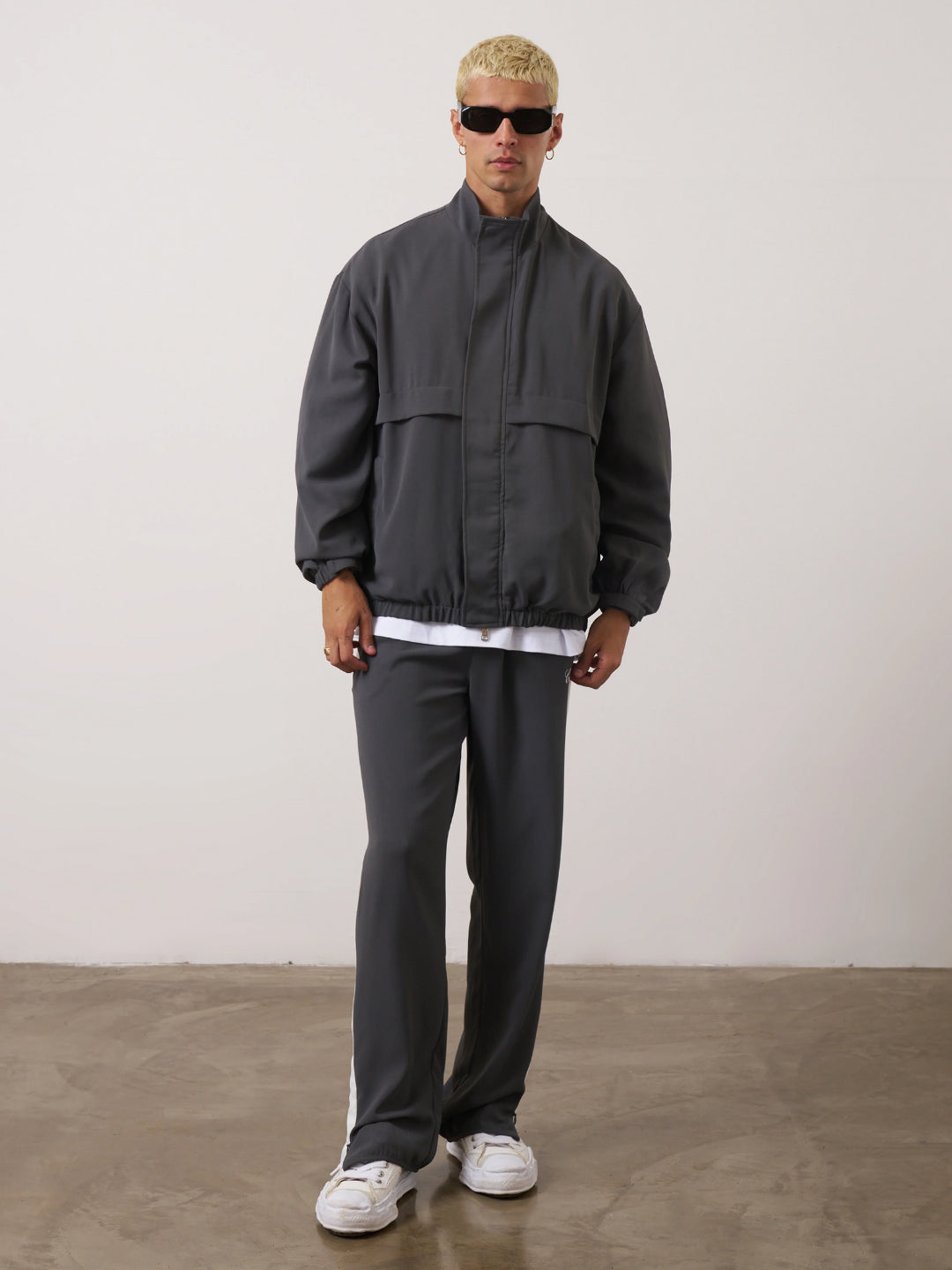 A man standing wearing a dark gray tracksuit, consisting of a jacket with a funnel neck and front pockets, and matching pants with an elasticated waistband.