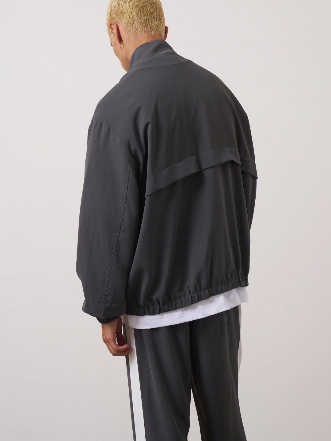 A man standing wearing a dark gray tracksuit, consisting of a jacket with a funnel neck and front pockets, and matching pants with an elasticated waistband.