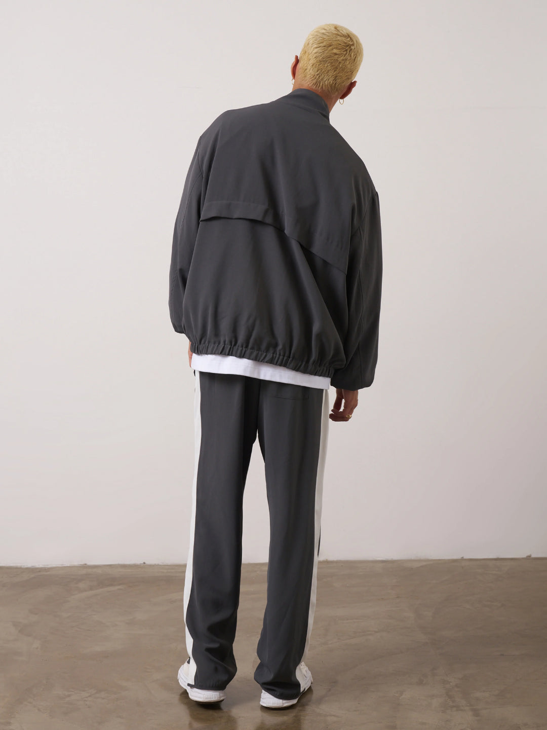 A man standing wearing a dark gray tracksuit, consisting of a jacket with a funnel neck and front pockets, and matching pants with an elasticated waistband.