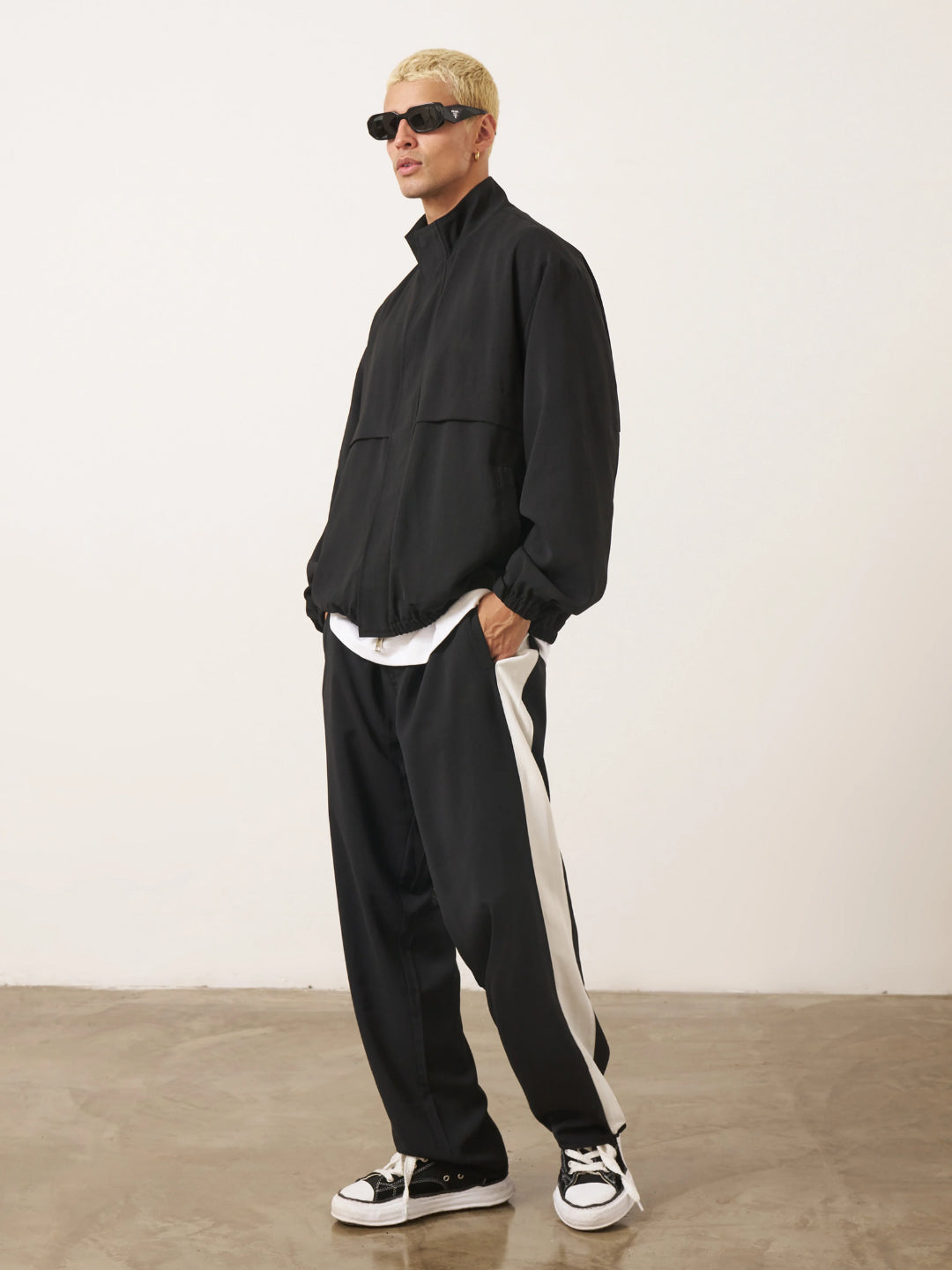 A man standing wearing a black tracksuit, with the jacket featuring a funnel neck and front pockets, and the pants having an elasticated waistband.