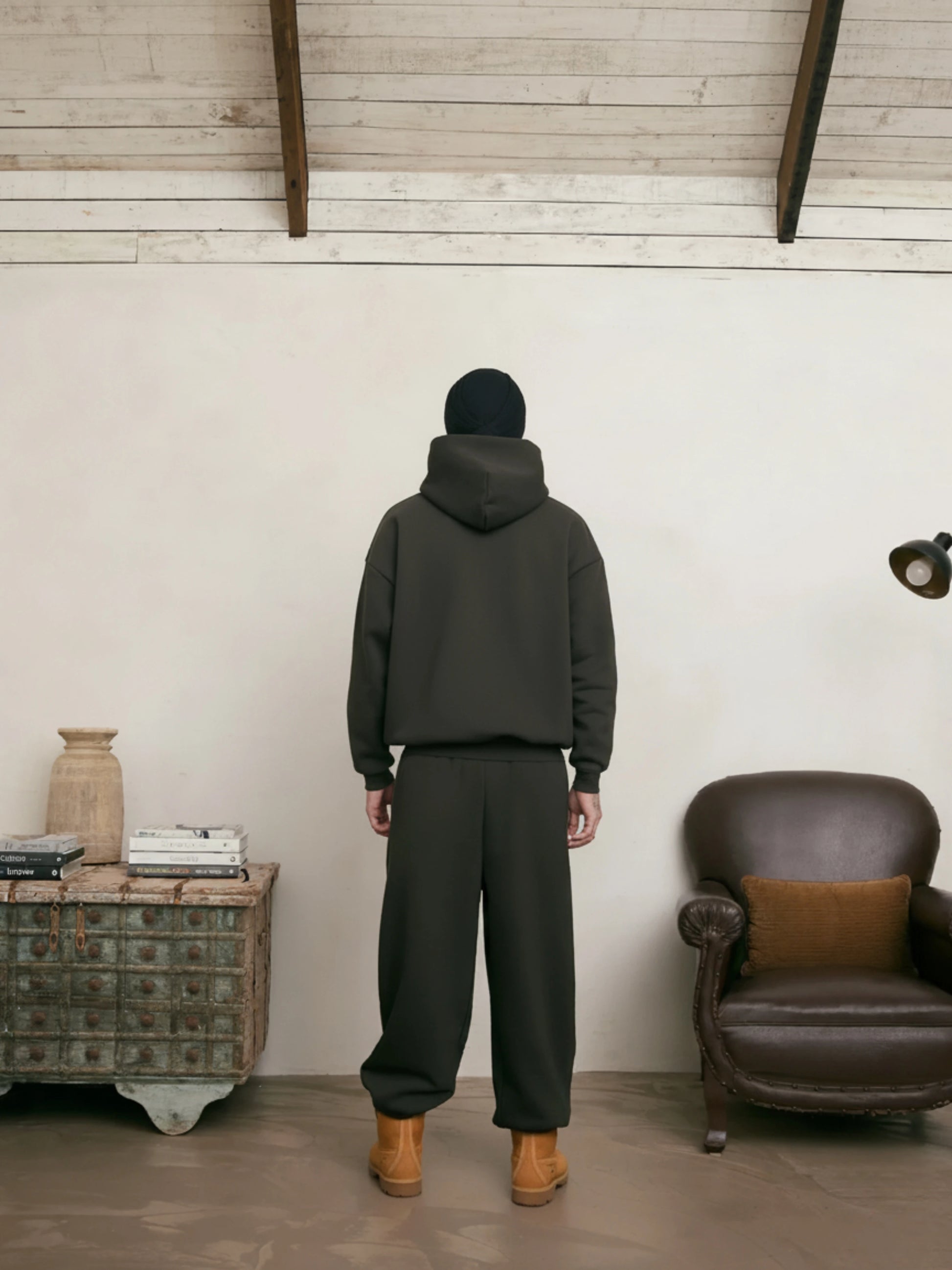 Person wearing a dark green hoodie and pants standing in a room with a brown leather chair and wooden side table.