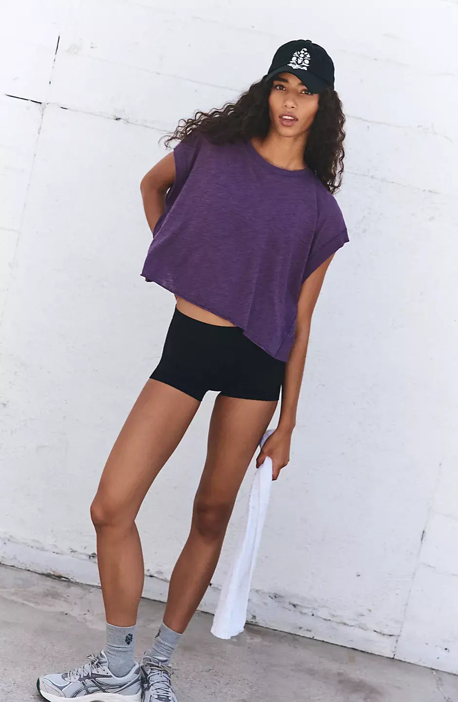 Woman wearing a purple t-shirt, black shorts, and a cap against a white wall.