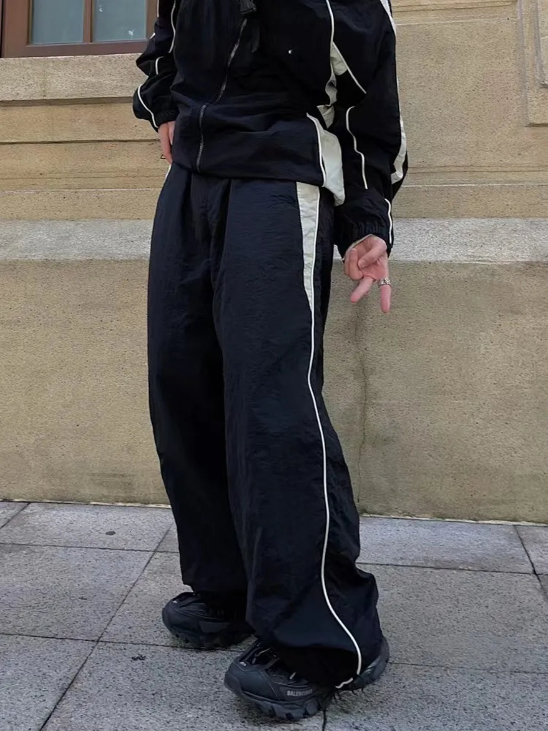 A person standing on a sidewalk wearing black trackpants with contrast white piping details and a black top, with hands in pockets.