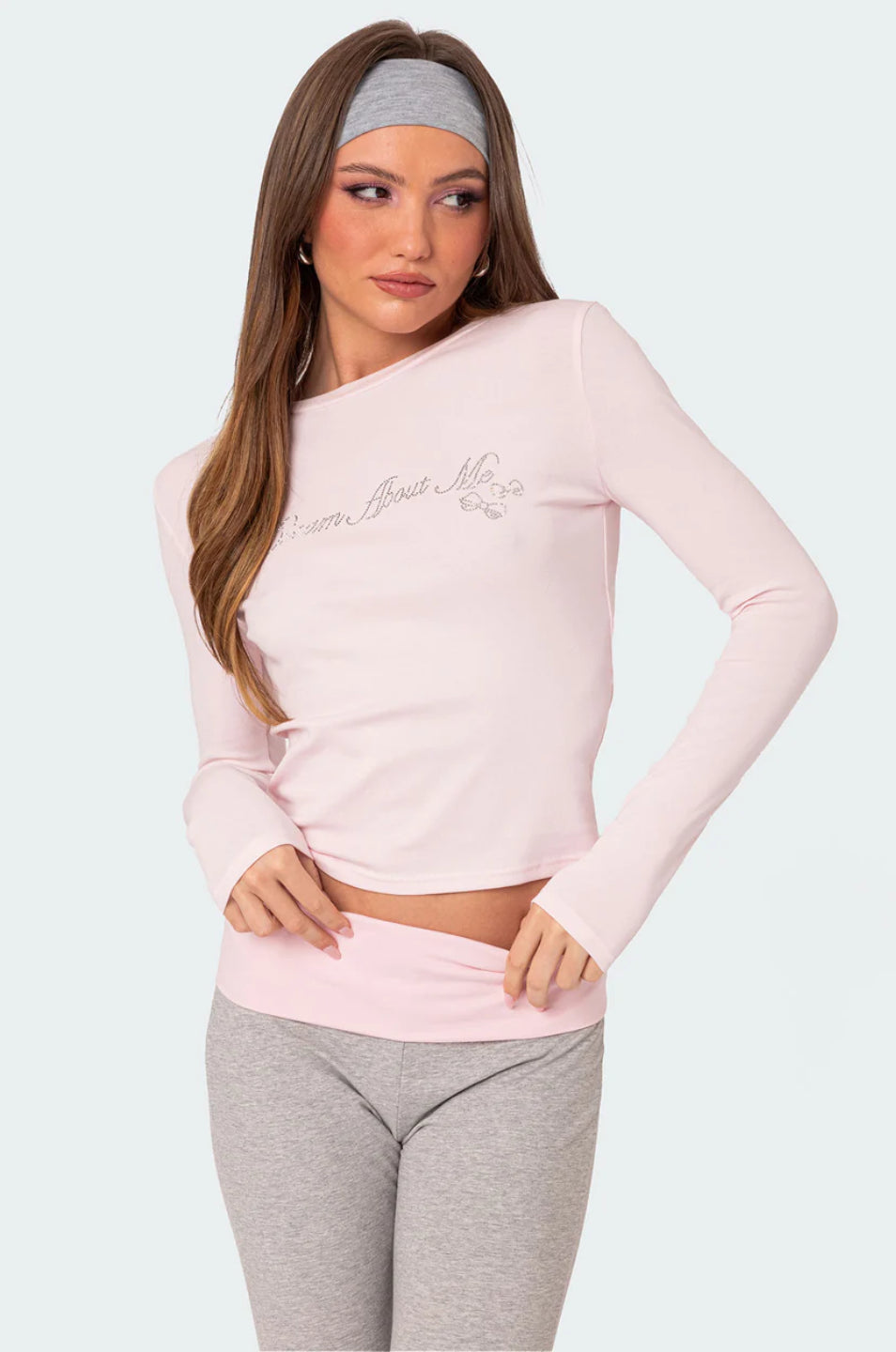 A female model wearing a pink rhinestone top with long sleeves, paired with gray flare leggings. She is also holding a small white purse.