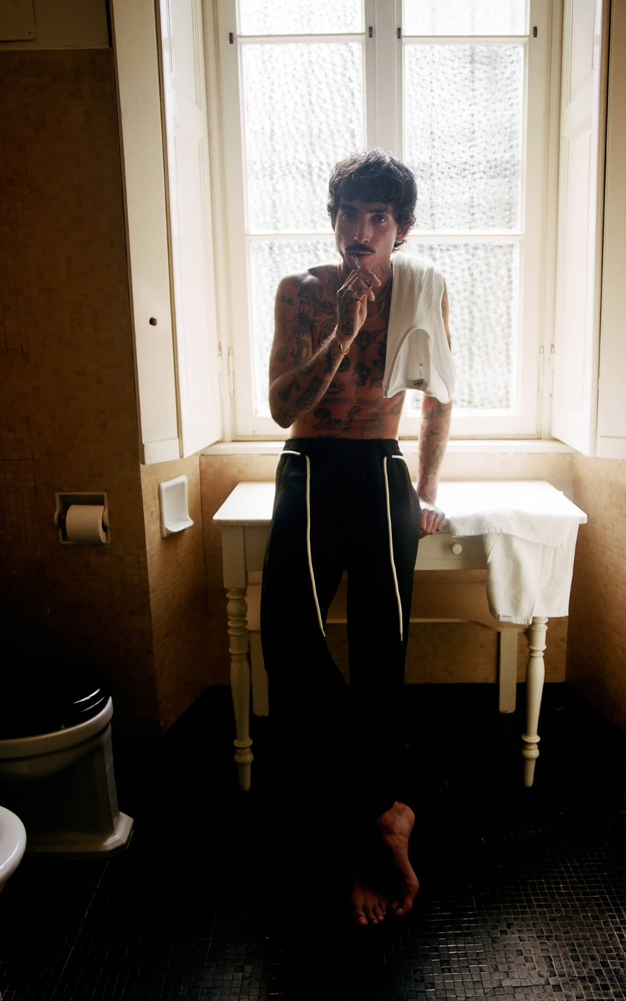 A person sitting on a bench in a room with a window, wearing trousers and a white shirt, with their back slightly turned to the camera.