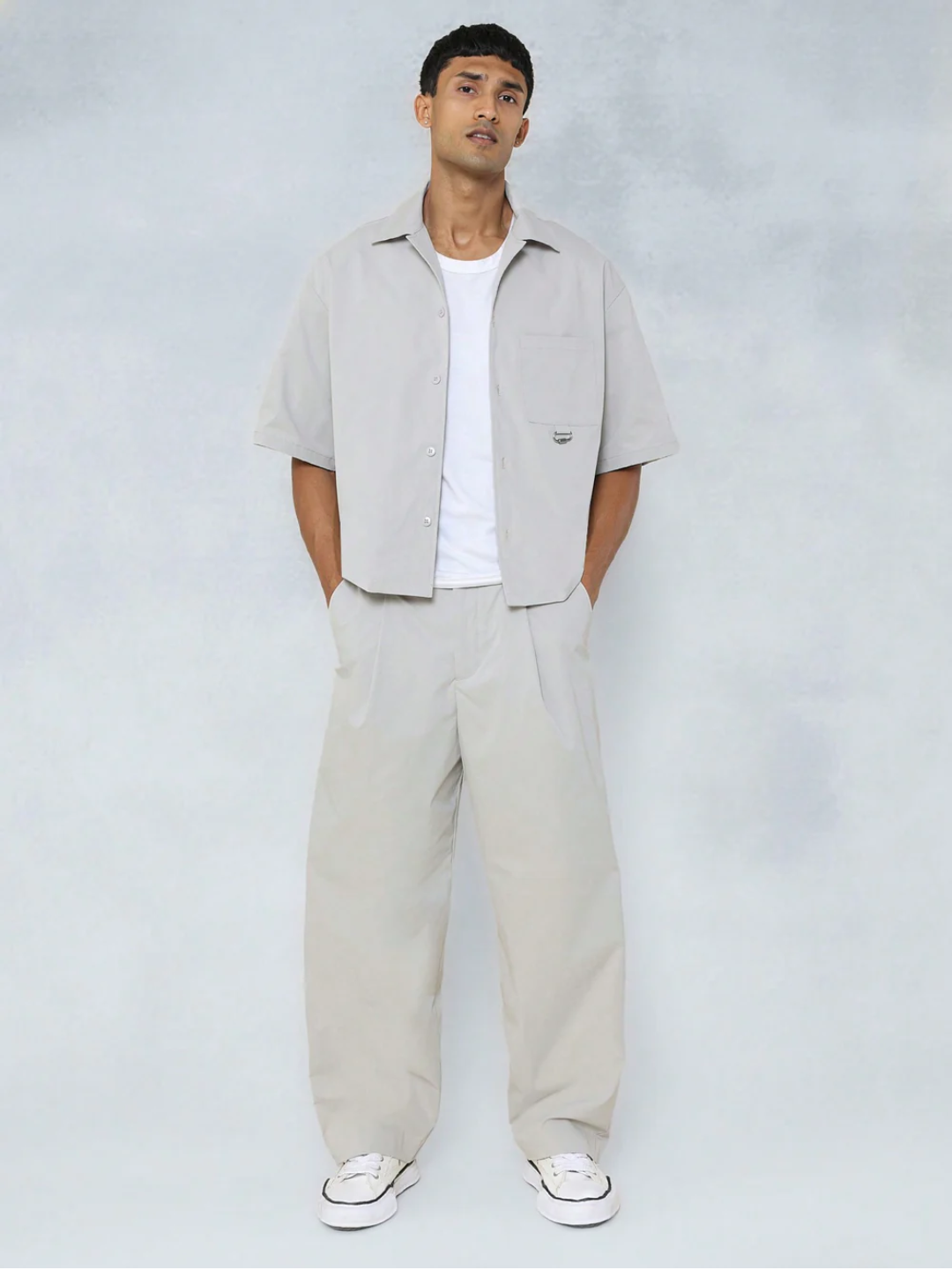A male model wearing a Grey crop fit shirt with a classic collar and drop shoulder sleeves, paired with beige loose fit pants.