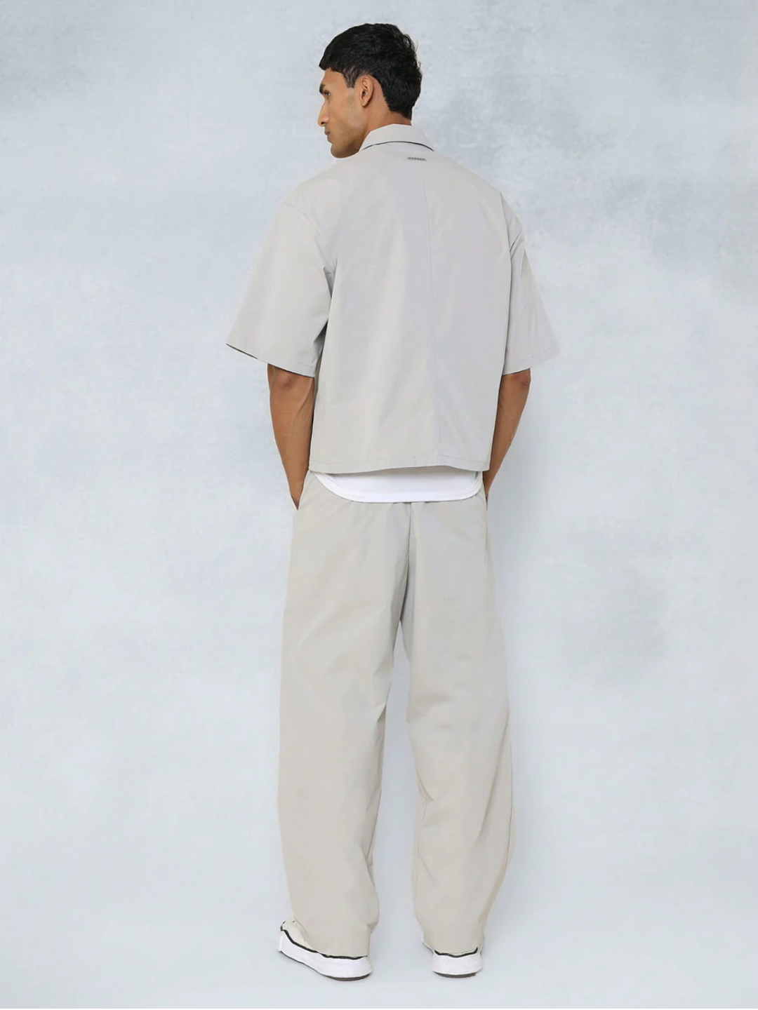 A male model wearing a Grey crop fit shirt with a classic collar and drop shoulder sleeves, paired with beige loose fit pants.