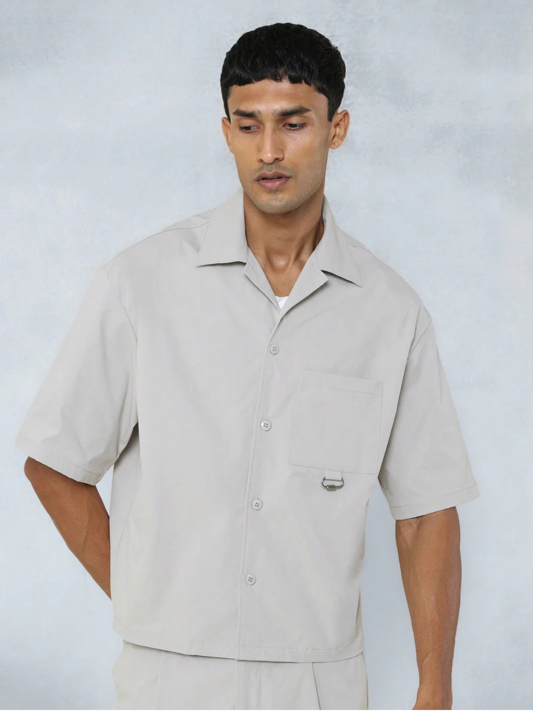 A male model wearing a Grey crop fit shirt with a classic collar and drop shoulder sleeves, paired with beige loose fit pants.