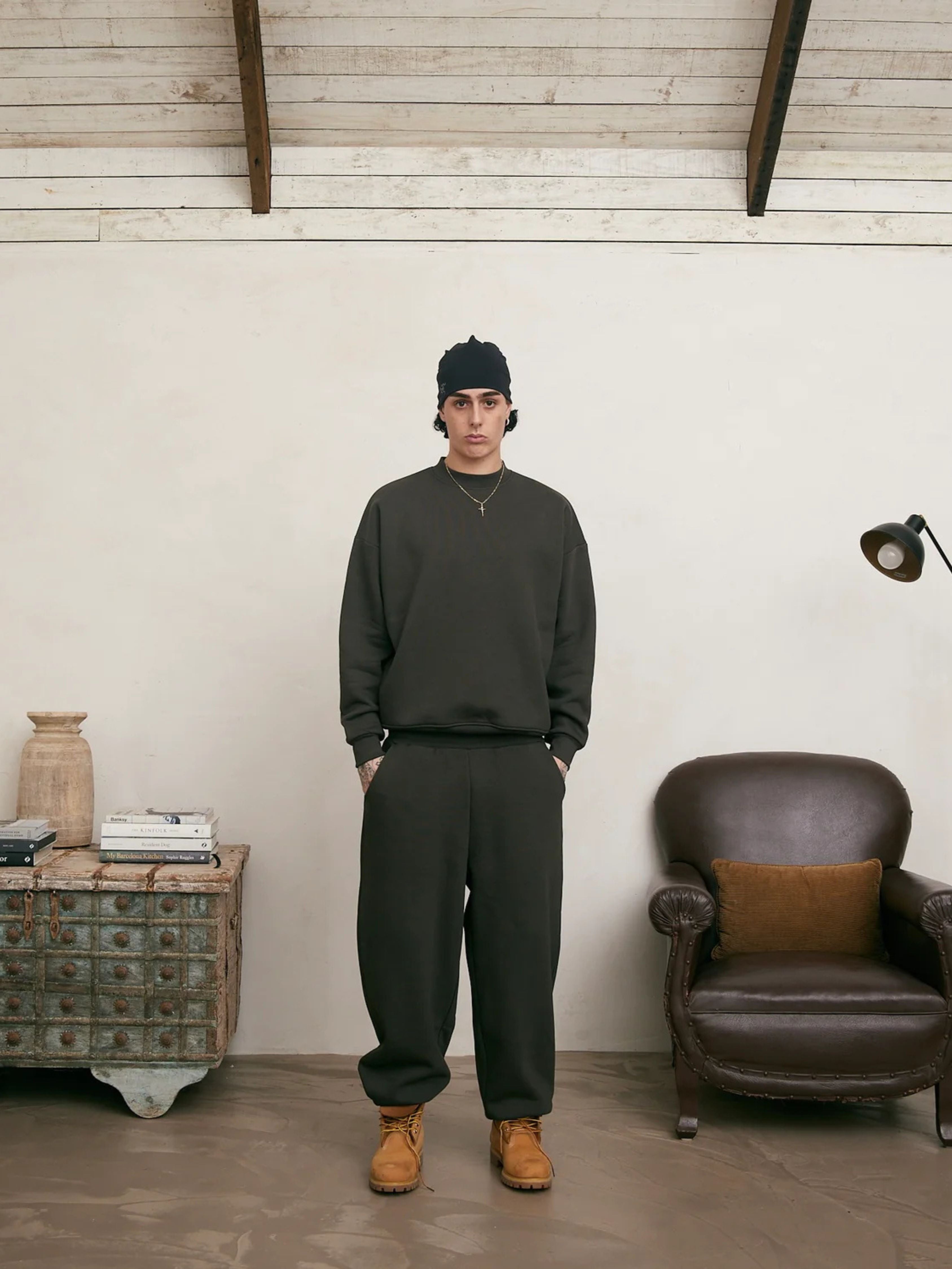 Person wearing a dark green outfit standing in a room with a brown armchair and wooden cabinet.