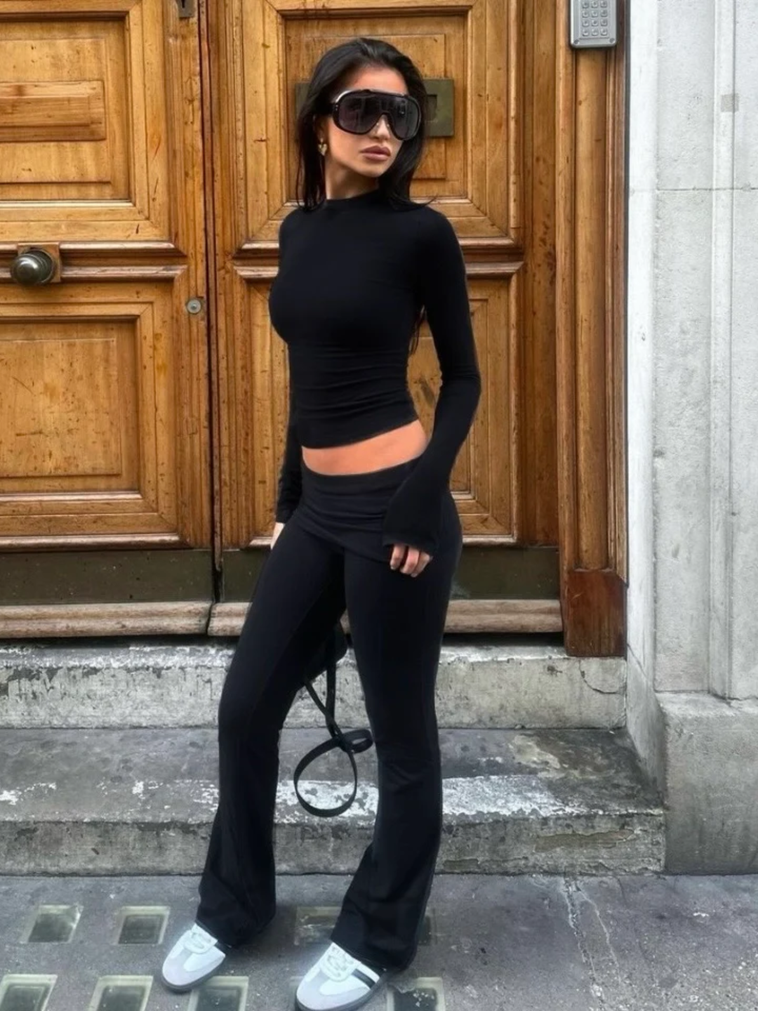 A woman wearing a black long-sleeve top and matching pants, standing in front of a wooden door.