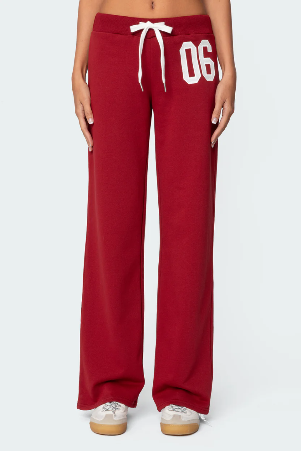 A female model wearing a white tank top and red lowrise trackpants with the number '06' printed on the left leg.