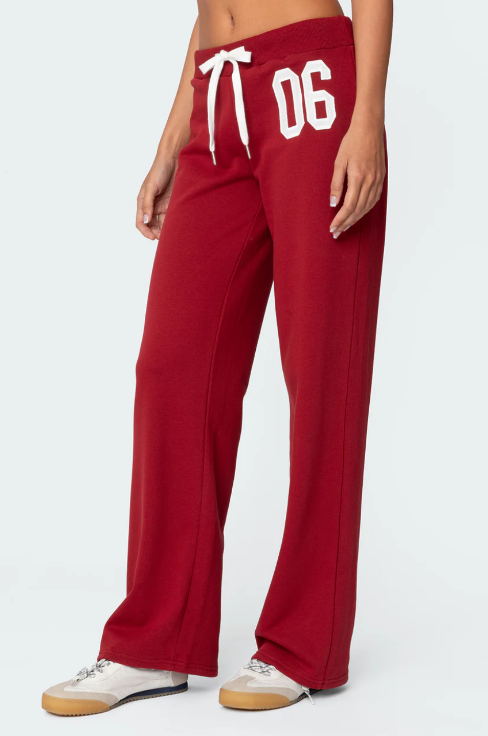 A female model wearing a white tank top and red lowrise trackpants with the number '06' printed on the left leg.