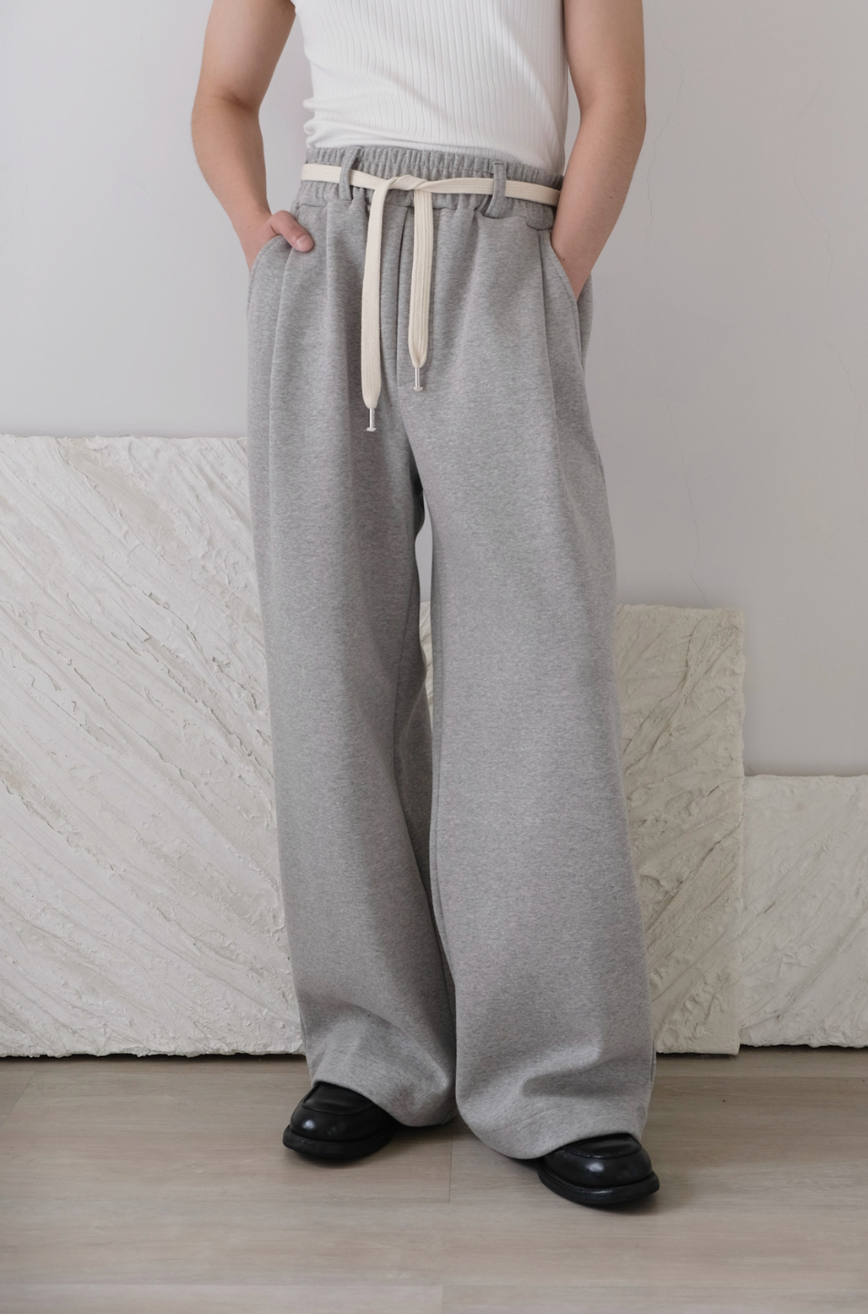 A person standing in a room wearing wide leg gray pants with a cotton belt, a white top, and black shoes.