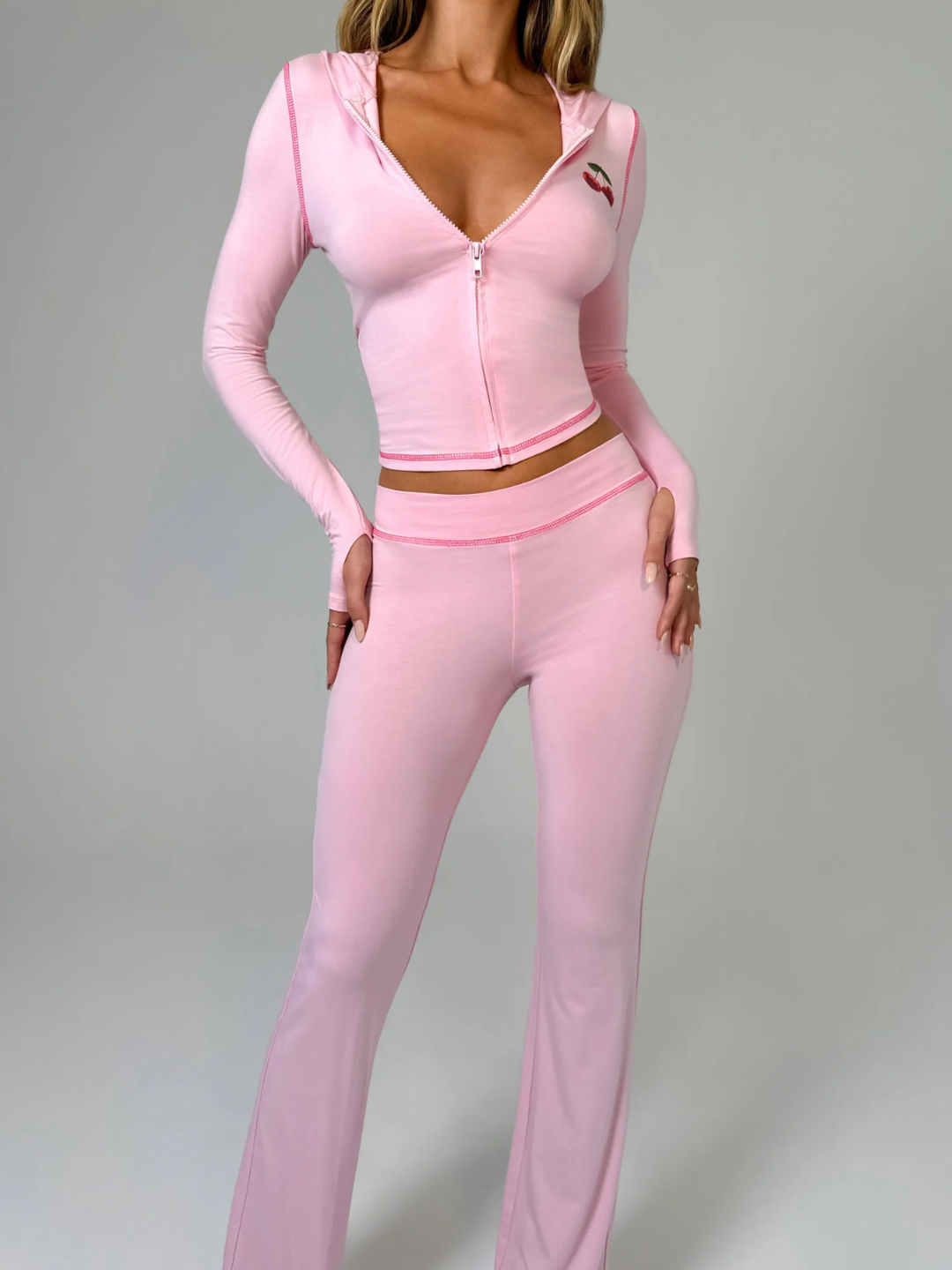 A female model wearing a pink long-sleeve hoodie and matching low-rise track pants with cherry placement prints and contrast stitching.