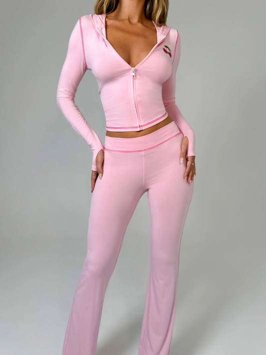 A female model wearing a pink long-sleeve hoodie and matching low-rise track pants with cherry placement prints and contrast stitching.