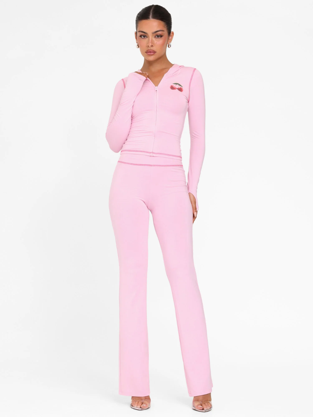 A female model wearing a pink long-sleeve hoodie and matching low-rise track pants with cherry placement prints and contrast stitching.