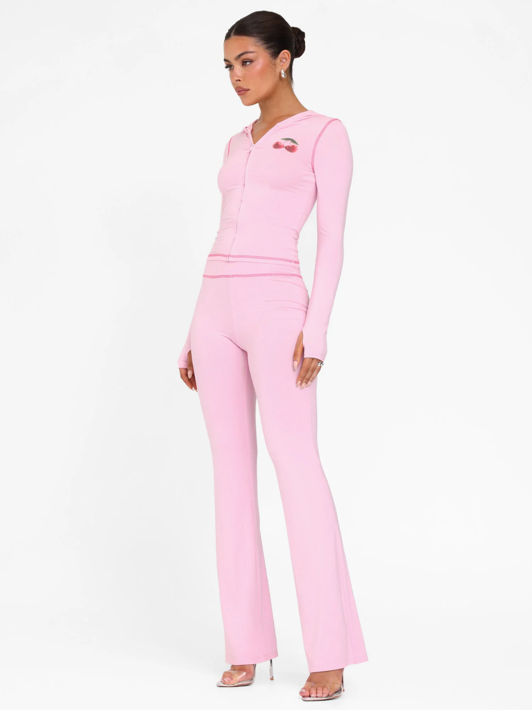 A female model wearing a pink long-sleeve hoodie and matching low-rise track pants with cherry placement prints and contrast stitching.
