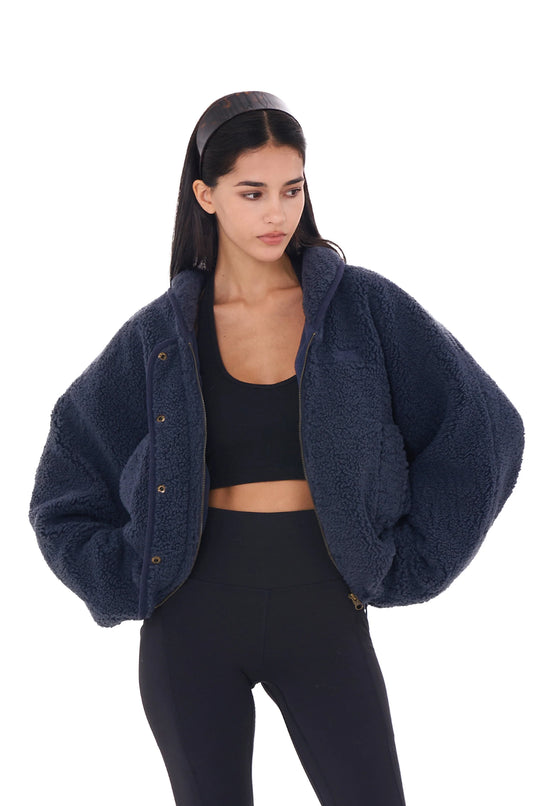A woman wearing a navy blue fleece zip jacket with a full-zip front, paired with black leggings. The jacket is double-sided with one side smooth and the other textured fleece.