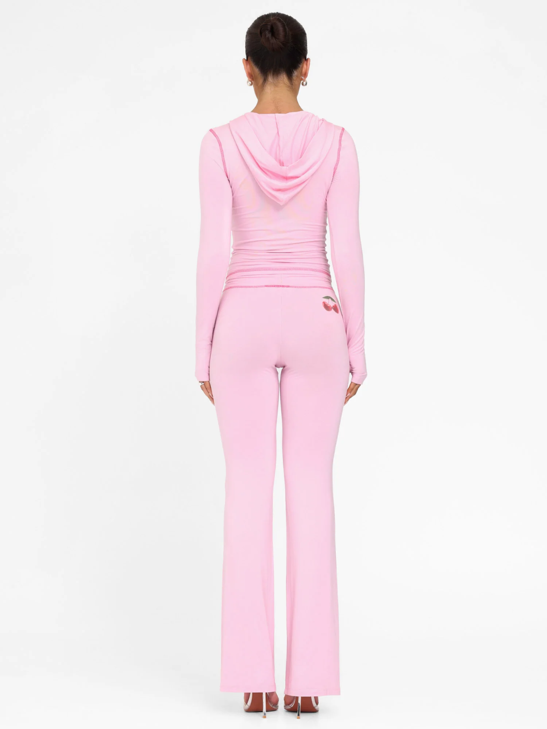 A female model wearing a pink long-sleeve hoodie and matching low-rise track pants with cherry placement prints and contrast stitching.