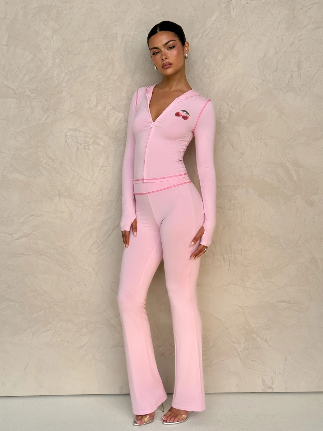 A female model wearing a pink long-sleeve hoodie and matching low-rise track pants with cherry placement prints and contrast stitching.