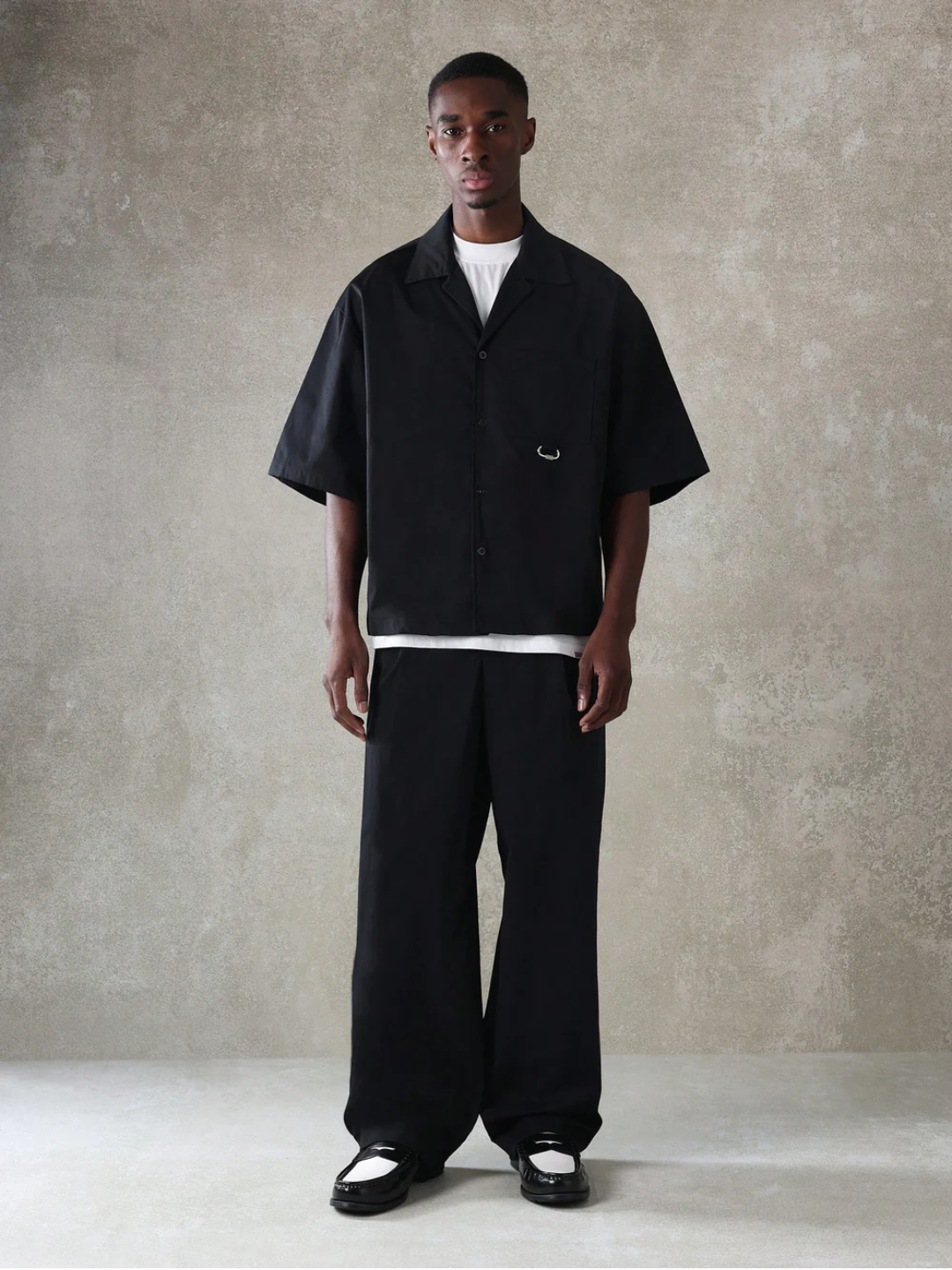 A male model wearing a Black crop fit shirt with a classic collar and drop shoulder sleeves, paired with beige loose fit pants.