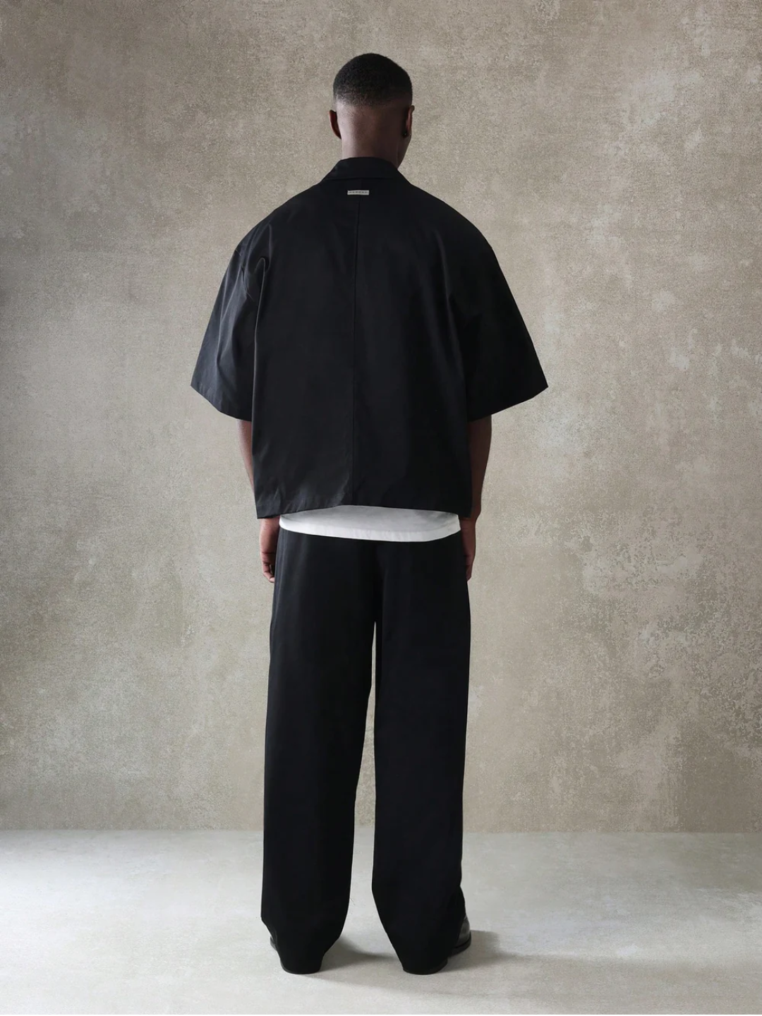 A male model wearing a Black crop fit shirt with a classic collar and drop shoulder sleeves, paired with beige loose fit pants.
