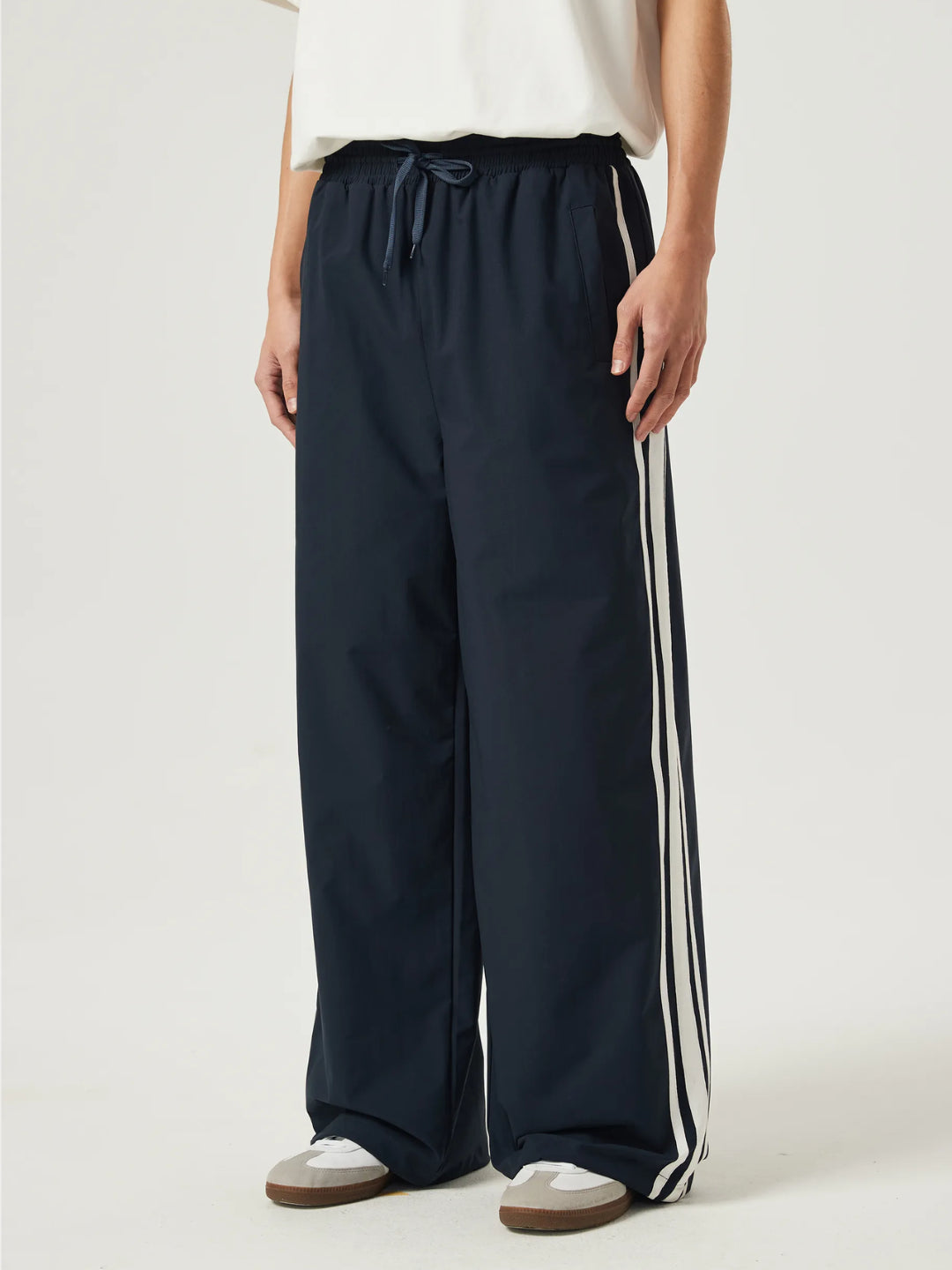A person standing wearing navy blue race track pants with white stripes on the side and an elasticated waistband with a drawstring.